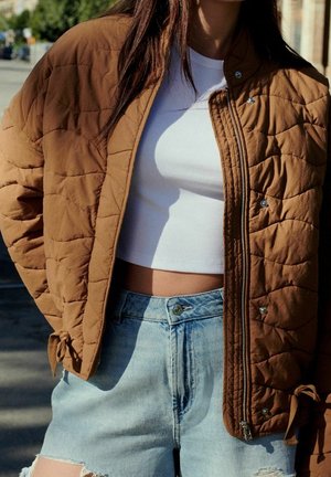 Veste matelassée marron avec boutons pression, associée à un crop top blanc et un short en jean clair. Caractérisée par des liens latéraux et une coupe décontractée.