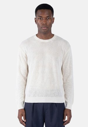 Uomo che indossa un maglione bianco a maniche lunghe di maglia e pantaloni blu scuro, in piedi su uno sfondo semplice grigio chiaro.