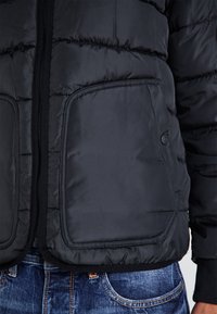 Giacca a vento nera con finitura testurizzata, dotata di una zip frontale e due grandi tasche con bottone in vita.