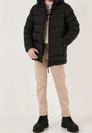 Person trägt eine schwarze Steppjacke, einen gemusterten Pullover, beige Hosen und schwarze Schnürstiefel und steht mit den Händen nahe den Taschen.