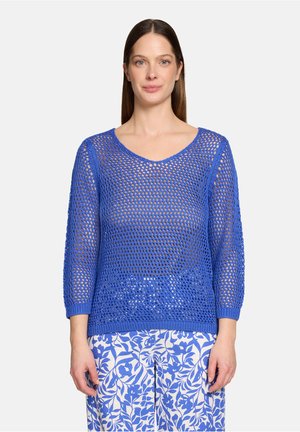 Femme portant un pull bleu en maille ajourée à manches trois-quarts et un pantalon blanc à motifs floraux bleus, debout devant un arrière-plan uni.