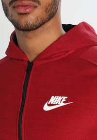 Červená mikina s kapitánem na zip vyrobená z texturované tkaniny, s černým interiérem, obepínající kapucí a výrazným bílým logem Nike.