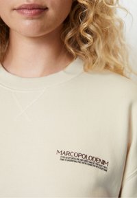 Žena s blond kudrnatými vlasy, oblečená v krémovém svetru s nápisem "MARCOPOLODENIM" a výšivkou s citátem na lesní téma na prsou.