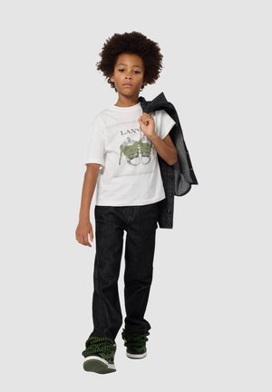 T-shirt blanche avec un graphisme de chaussures vertes, jean noir et baskets avec accents verts. Le modèle porte une veste grise sur l'épaule.