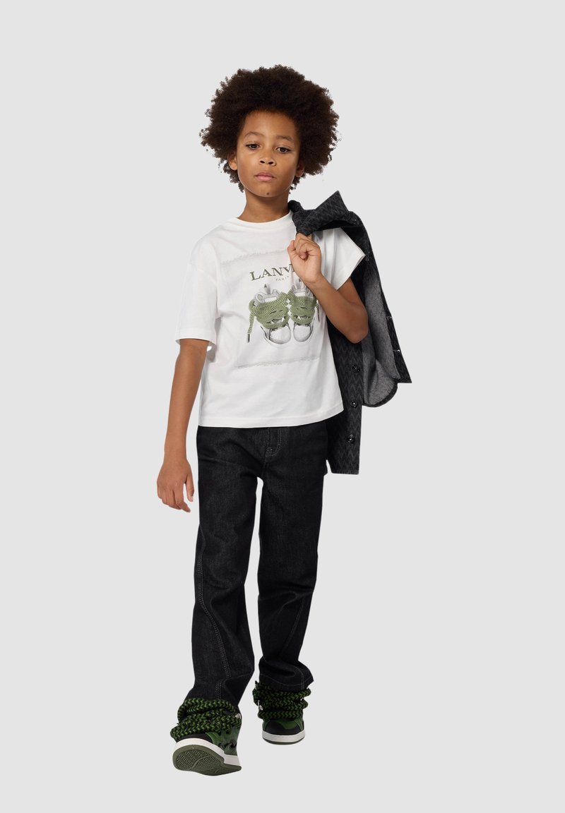 Biała koszulka z grafiką zielonych butów, czarne dżinsy i sneakersy z zielonymi akcentami. Model trzyma szary żakiet na ramieniu.