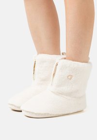 Cozy white boot slippers with a soft, plush exterior, side zippers, and a round logo accent near the top. Features a flat, stitched sole.