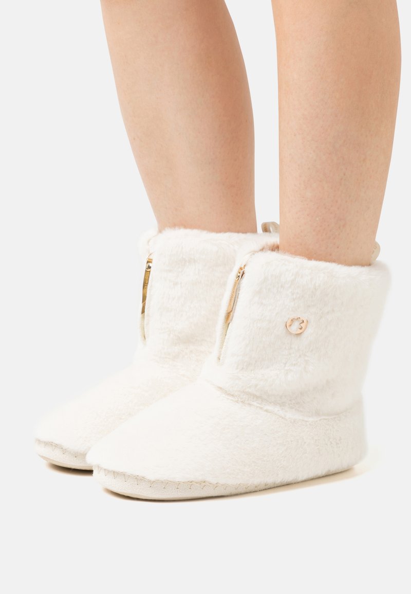 Cozy white boot slippers with a soft, plush exterior, side zippers, and a round logo accent near the top. Features a flat, stitched sole.