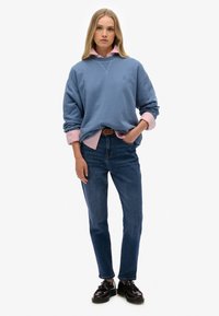 Lichtblauwe oversized sweatshirt met ronde hals, gelaagd over een roze overhemd met kraag. Gepaard met donkerblauwe denim jeans en zwarte schoenen.
