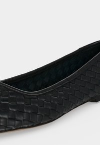 Chaussure plate noire avec texture en cuir tissé et bout arrondi, présentée légèrement en angle sur un fond clair.