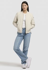 Quiltad ljusbeige bomberjacka med rund hals och sidodragkedjor, ihop med blå raka jeans och vita sneakers.