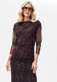 Brązowa sukienka w wzór skóry węża z przezroczystymi, długimi rękawami, marszczeniami i dopasowanym krojem. Uzupełniona dużymi brązowymi okularami przeciwsłonecznymi.