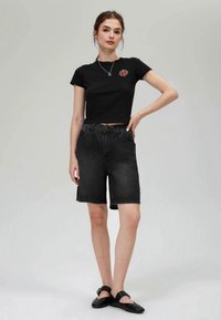 T-shirt noir court avec un graphique rouge sur la poitrine, associé à un short en denim foncé. Le mannequin porte des chaussures plates noires et un collier simple.