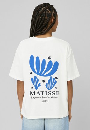Femme aux cheveux tressés portant un T-shirt blanc avec des formes abstraites bleues et le texte noir « MATISSE La perruche et la sirène (1958) » au dos.