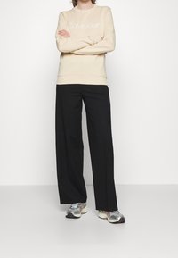 Persona in piedi con le braccia incrociate, indossa un felpa beige di Calvin Klein, pantaloni neri a gamba larga e scarpe da corsa grigie.