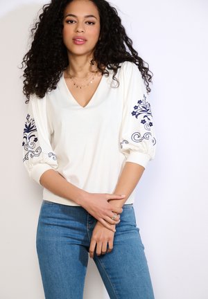 Žena s kudrnatými vlasy, která nosí bílou blůzu s modrou květinovou výšivkou na nafouknutých rukávech a modré džíny, stojící před jednobarevným pozadím.