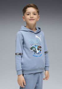 Ragazzo giovane che indossa una felpa leggera blu di Puma e pantaloni, sorridendo leggermente, con grafiche di monster truck e testo sulla parte anteriore della felpa.
