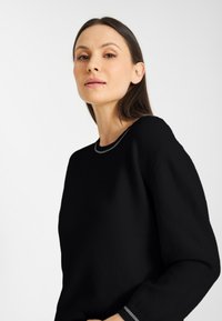Vrouw met lang, donker haar, die een zwarte trui draagt met een grijze bies op de kraag en manchetten, poserend tegen een eenvoudige witte achtergrond.