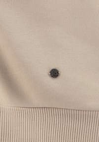 Un tessuto beige a coste con una texture liscia presenta un dettaglio in metallo circolare con incisione "Ky", posizionato in modo sobrio sul tessuto.