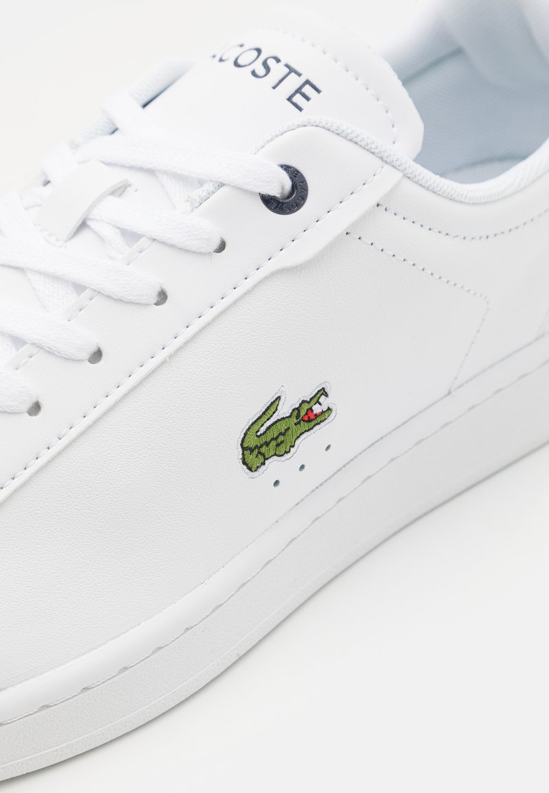 Baskets blanches avec une tige en cuir synthétique lisse, fermeture à lacets, logo de crocodile vert et perforations discrètes sur le côté.