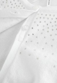 Detail bílého pleteného kardiganu s malými stříbrnými nýty zdobícími látku a bílým knoflíkem na předním rozparku.