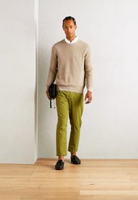 Pull en tricot beige, chemise blanche à col, chino vert et mocassins noirs. La personne porte un sac noir et se tient sur un sol en bois.