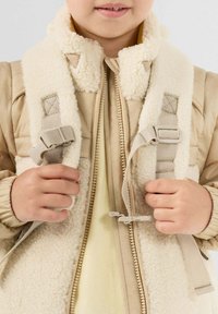 Kind draagt een beige jas met fleece voering, houdt riemen van een beige rugzak vast, met rits gedeeltelijk zichtbaar aan de voorkant van de jas.