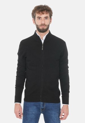 Uomo con capelli corti ricci e barba, che indossa un maglione nero con zip sopra una camicia bianca e jeans blu, in piedi su uno sfondo bianco.
