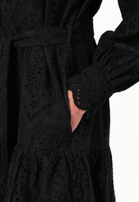 Svart kjole med intrikate broderte mønstre, teksturert stoff, innsvingt midje med belte og puffede ermer med bølgete mansjetter.