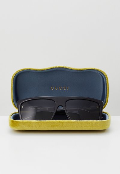 Gucci päikeseprillid mustas raamis, esitatud kollases ja sinises sametkarbis. Karbi serv on laineline ja sellel on reljeefne logo.