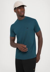 Pasztell zöld crewneck póló, puha anyagból készült, diszkrét fekete logóval a bal mellen. A modellen fekete nadrág és bézs sapka van.