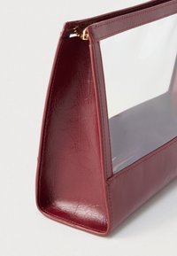 Μπορντό τσάντα με γυαλιστερό φινίρισμα, διαφανή πλαστική πλευρά, ορθογώνιο σχήμα, λεία υφή και χρυσό φερμουάρ στην κορυφή.