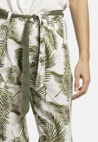 Pantalones de lino blancos con patrón de hojas de palma verdes, que cuentan con un lazo de tela a juego en la cintura y bolsillos laterales. Textura suave.