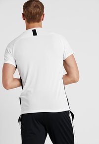 Camiseta atlética blanca con un diseño texturizado, mangas cortas y detalles negros en los lados. La vista trasera muestra un corte recto y una etiqueta.