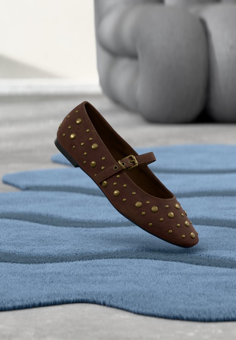 Chaussure Mary Jane plate marron avec des clous décoratifs dorés et une boucle, posée sur un tapis bleu texturé avec un canapé gris en arrière-plan.