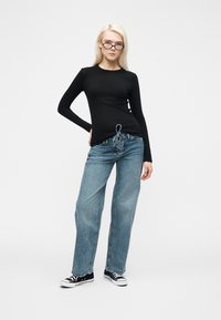 Νεαρή γυναίκα που φορά στενά μαύρα γυαλιά, μαύρο μακρυμάνικο μπλουζάκι, μπλε τζιν με κορδόνια και μαύρα αθλητικά παπούτσια, στέκεται με το ένα χέρι στη μέση.