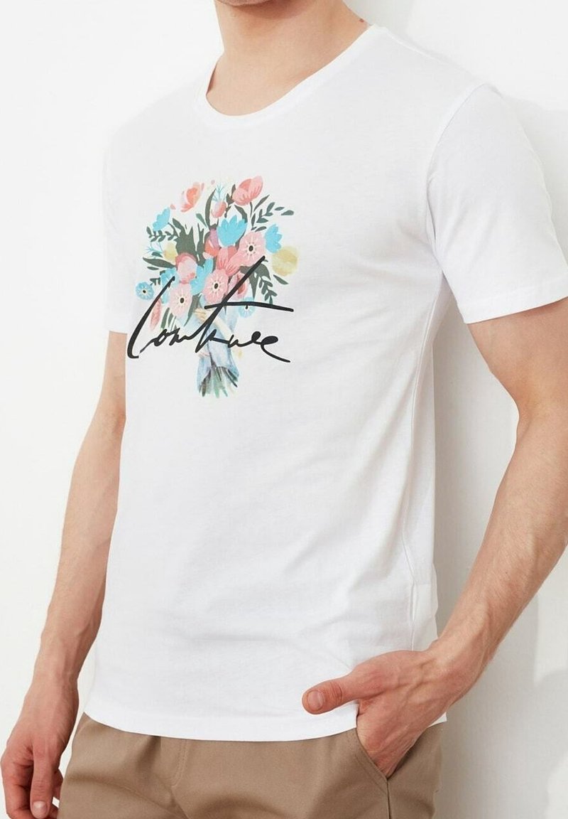 T-shirt blanc à manches courtes avec un bouquet floral coloré et le texte en cursive noire "couture", porté par une personne ayant une main dans la poche d'un pantalon beige.