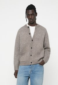 Cardigan tricotat de culoare maro deschis, cu guler rotund, butoni frontal și două buzunare. Material texturat și manșete cu nervuri pentru o ajustare confortabilă.