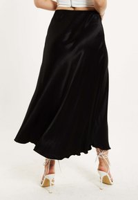 Black satin maxi skirt with a flowing design and a wavy hem. Features a fitted waistband, worn with white strappy high-heeled sandals.