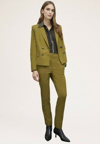 Completo su misura verde oliva con giacca doppiopetto e bottoni neri, abbinato a una camicia nera a trama. Indossato con stivaletti neri alla caviglia.