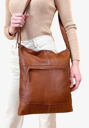 Brun læder skuldertaske med et slouchy design, prydet med en frontlomme og justerbar rem, holdt af en person i afslappet tøj.