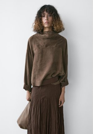 Donna con capelli ricci che indossa una maglia larga in camoscio marrone e una lunga gonna marrone plissettata, che tiene una borsa beige contro un muro bianco semplice.