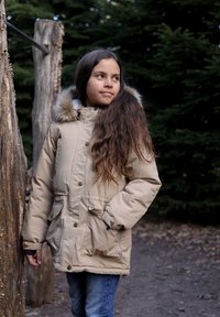 Beige Winterjacke mit pelzgefütterter Kapuze, ausgestattet mit Reißverschluss und Druckknopfverschluss, großen Fronttaschen und elastischen Bündchen. Im Freien.