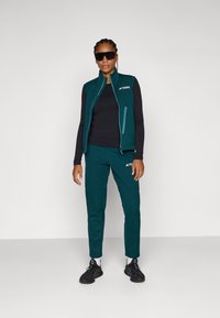 Türkisfarbener Sportweste mit Reißverschlusstaschen, kombiniert mit schwarzem Langarmoberteil und türkisfarbenen Hosen. Das Modell trägt schwarze Sonnenbrillen und Schuhe.