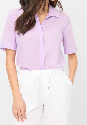 Kvinde iført lyse lilla kortærmet skjorte med knapper, stoppet ned i hvide bukser med snøre, stående med hænderne i lommerne.