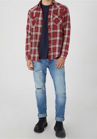 Chemise en flanelle à carreaux rouges et blancs avec deux poches poitrine, portée sur un t-shirt bleu marine. Jeans bleu clair déchirés et bottes noires à lacets.