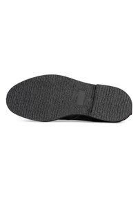 Suela de zapato de goma negra con superficie texturizada, que presenta surcos para tracción y una forma plana y redondeada. Incluye una pequeña etiqueta rectangular de marca.