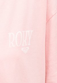 Ανοιχτό ροζ ύφασμα με λευκό κείμενο "ROXY" και ένα μικρό λογότυπο σε σχήμα καρδιάς κάτω από το κείμενο στην αριστερή πλευρά.