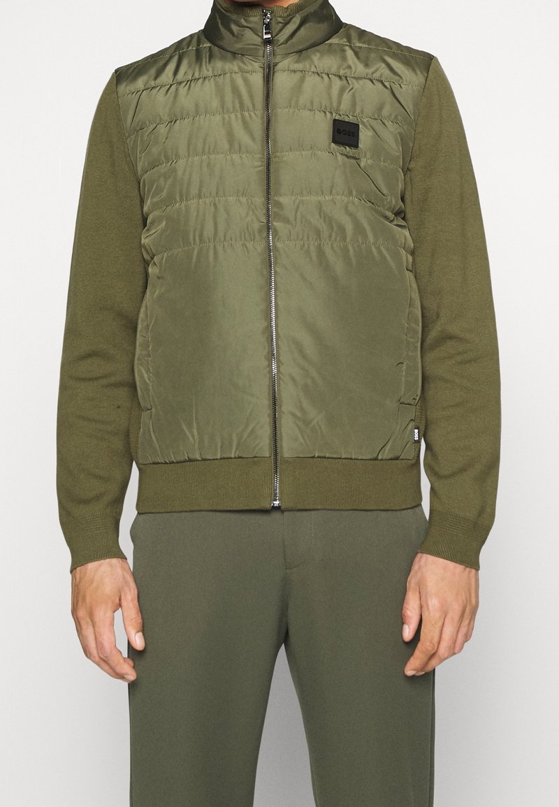 Homme portant une veste matelassée zippée vert olive avec manches tricotées assorties et pantalon, debout, les bras relâchés le long du corps.