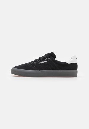 Baskets basses Adidas noires avec semelle grise, lacets noirs et patch blanc au talon avec le logo Adidas, vues de côté sur fond blanc.
