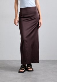 Femeie îmbrăcată cu un top alb fără mâneci, fustă lungă de un brun închis și sandale negre cu cataramă, stând pe un podea gri, în fața unui perete simplu.